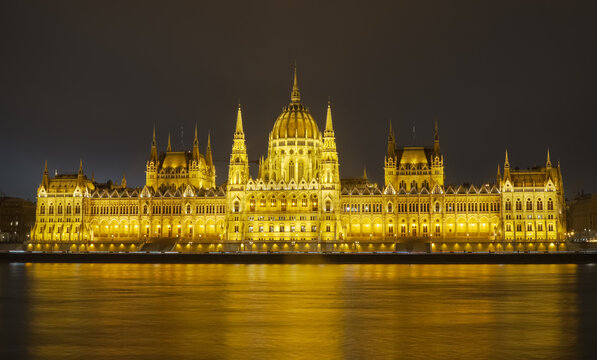 View of the illuminated Hungarian Parliament Building beside the Danube River in Budapest, Hungary, at night, with golden reflections and its historic Gothic Revival facade in winter.