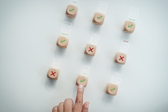 Hand touching wooden block with green check mark icon among cross mark blocks with digital document approval interface overlay on white desk, concept of quality control and compliance.