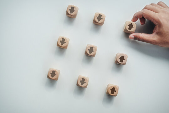 Hand placing wooden block with upward arrow icon among downward arrow blocks on white desk, concept of business growth, market recovery, success trend, and positive direction.