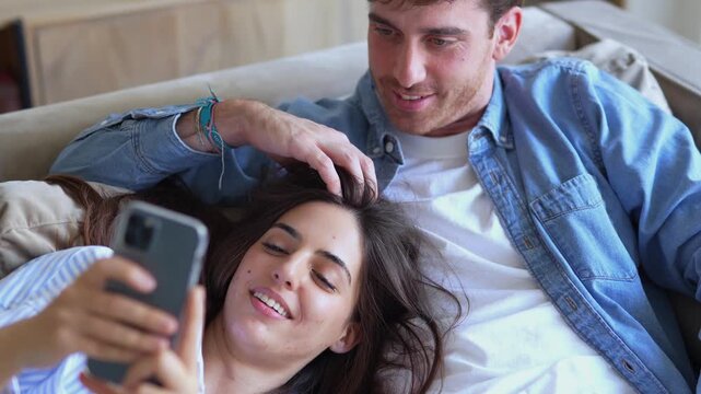 Young happy couple spending quality time together, taking a selfie with a smartphone while relaxing on the sofa. The smiling man is affectionately stroking his girlfriend's hair