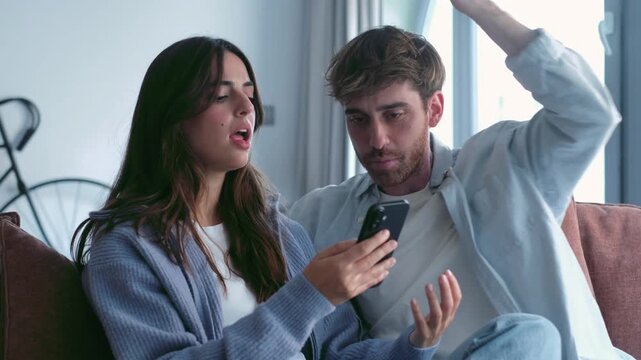 Frustrated young man and woman arguing about a message on a mobile phone while sitting on the couch in their living room, experiencing jealousy, mistrust, and relationship problems at home