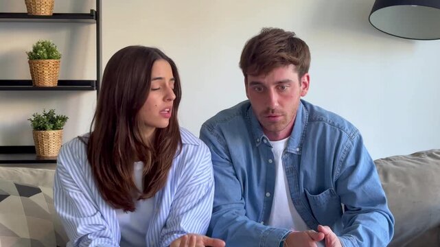 Young married couple sitting on the couch discussing relationship issues, gesturing and talking with worried expressions in a modern living room while trying to resolve their conflict