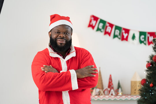 Portrait of African American father in red Santa hat and red Santa Claus costume celebrates Christmas in festive living room near decorated tree and festive decorations