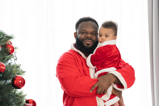 African American father in red Santa Claus costume celebrates Christmas during holding  little baby boy in festive living room near decorated tree and festive decorations
