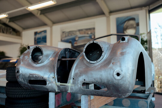 View of a bare metal classic car body under restoration in a workshop with stacked tires and automotive posters in the background in Koudekerke, Zeeland, Netherlands.