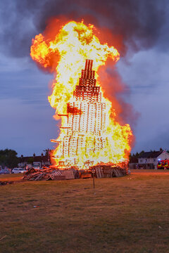 View of a large bonfire made of stacked wooden pallets burning brightly with intense flames and smoke in a field at dusk in Larne, Northern Ireland, United Kingdom.