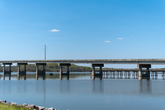 View of a concrete bridge spanning across a calm lake with a small boat visible near the pillars under a clear blue sky in Camden, Alabama, United States.