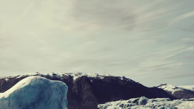 Huge glacier ice block resting near dark rocky mountains covered with snow under an overcast sky during daytime