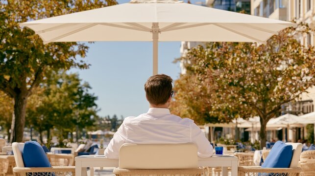 Man in white shirt sitting alone at outdoor caf&eacute; table under large umbrella