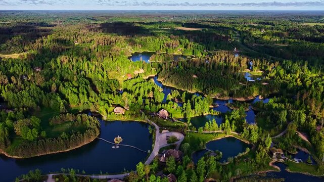 Aerial view of a lush green forest landscape featuring multiple small lakes, winding paths, and scattered wooden cabins in Latvia.