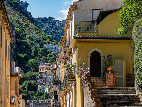 Historic street scene featuring yellow buildings, stone steps, and mountain slopes in Taormina, Sicily, Italy.