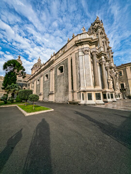 Vertical composition highlighting the carved marble columns, volcanic basalt masonry wall, dome silhouette, and cloud formations of Catania Cathedral.