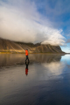 View of a person in an orange jacket walking on the wet, reflective black sand beach at Vestrahorn with dramatic mountains partially covered by clouds, Iceland.