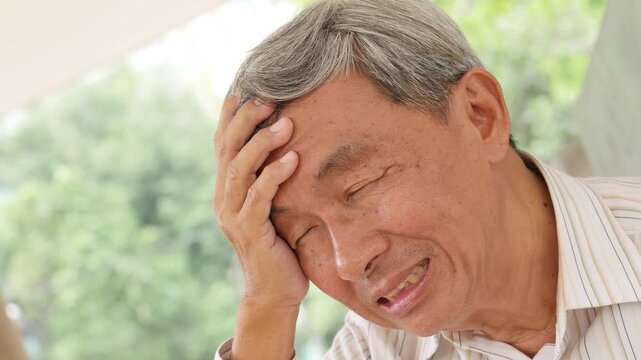 elderly east asian male in his 60s with headache pain at home in daylight, looking worried and sad