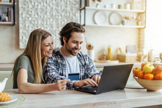 Smiling Young Couple, Caucasian And Asian, Working Together On Laptop At Kitchen Table, Collaborating And Enjoying Each Other's Company In Cozy Home Setting. Family Concept.