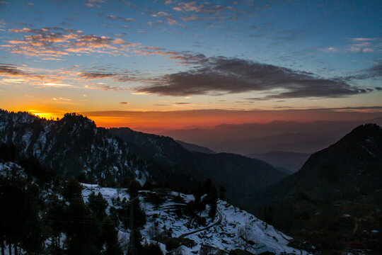 View of Malam Jabba mountains covered in snow during a vibrant sunset with orange clouds and dark silhouettes of pine trees in Swat, Khyber Pakhtunkhwa, Pakistan.