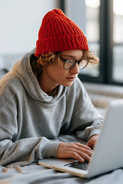 Young boy wearing a red beanie and glasses lying down while using a laptop computer.