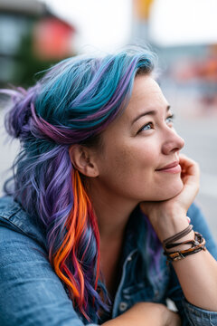 Smiling young woman with creative blue, purple, and orange dyed hair looking up thoughtfully outdoors.