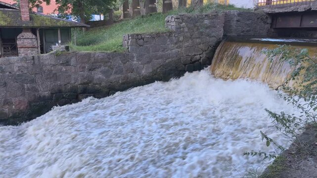 A wide view shows water rushing over an urban weir and churning below a nearby bridge. 