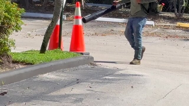 Landscaping worker using a gas-powered backpack leaf blower to clean a parking lot pavement