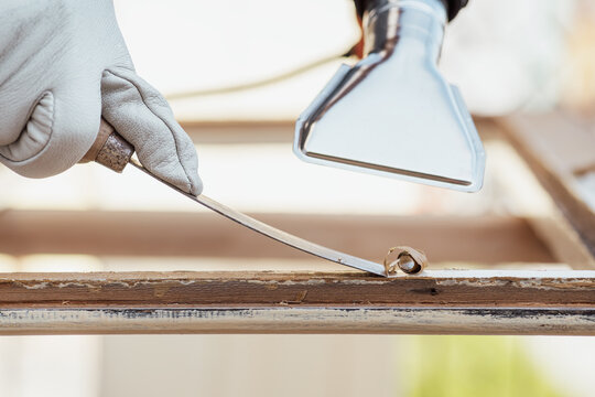 Adult carpenter wearing protective leather gloves, using a heat gun and a wood scraper to remove paint from an old wooden window. Construction industry, carpentry. Restoration. 