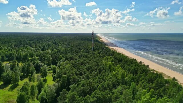 Aerial view of a communication tower rising above a dense green forest bordering a sandy beach and the Baltic Sea in Latvia.