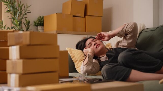 Exhausted Asian female entrepreneur lying on couch resting hand on forehead, overwhelmed by stress and workload of online business.