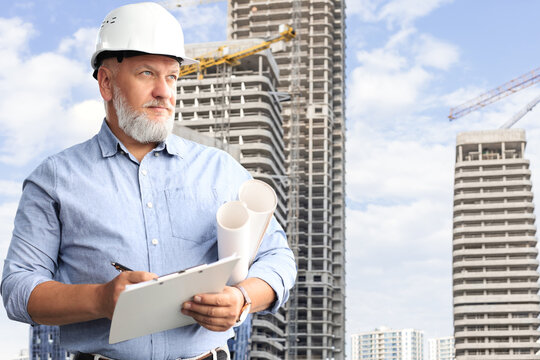 Confident mature business man in hardhat holding blueprint of building to skyscraper construction, buildings under construction in the background.