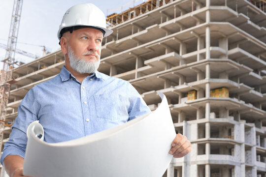 Confident mature architect in corporate suit and hardhat holding a blueprint and looking at it.