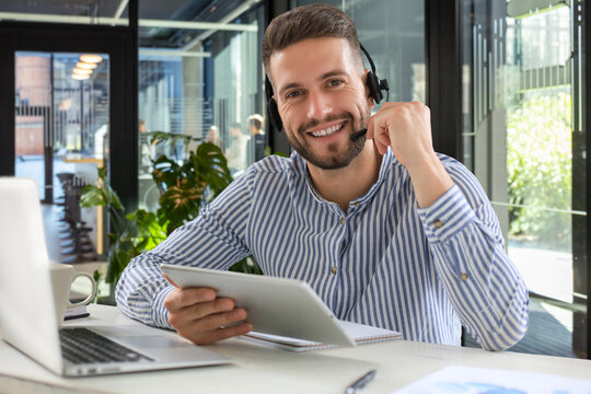 Cheerful young support phone male operator in headset, at workplace while using digital tablet in office