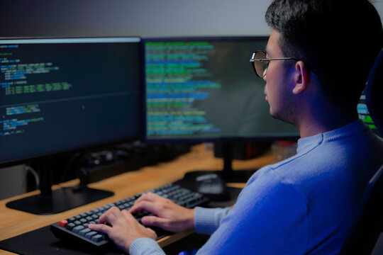 Computer programmer works at a multi monitor workstation in a dark office, reviewing programming code for software development, technical analysis, and system monitoring.