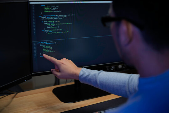 Computer programmer works at a multi monitor workstation in a dark office, reviewing programming code for software development, technical analysis, and system monitoring.