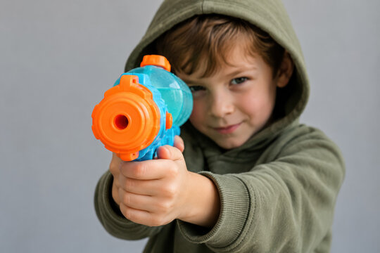 Boy in a hooded sweatshirt aims a colorful toy water gun toward the camera against a plain gray background.