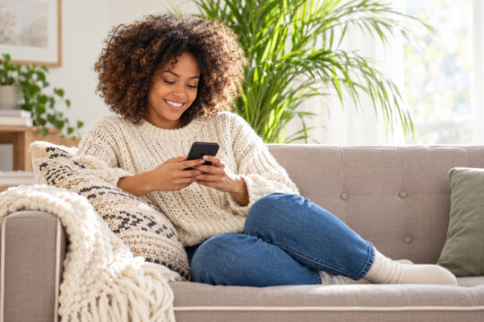 Young woman smiles while using a smartphone on a comfortable sofa at home. Bright indoor lifestyle setting with plants, cushions, and a knitted blanket.