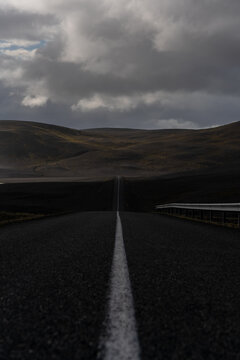 View of a long asphalt road stretching through the vast volcanic landscape of the Southern Region under a dramatic cloudy sky in Iceland.
