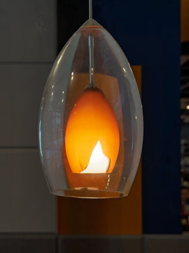 Pendant lamp with a clear outer glass shade surrounding an amber inner globe, a flame-shaped opening revealing the bright bulb, against blue, gray and orange panels.