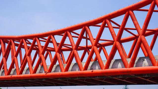 Red steel truss elevated railway bridge at Simpang Joglo, Surakarta, Indonesia. Modern geometric infrastructure architecture against a bright blue sky, showcasing urban transport design.