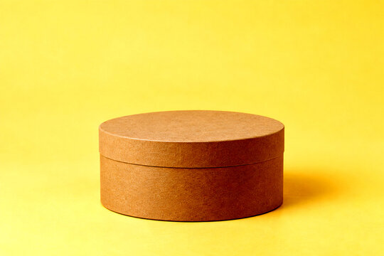 Round cardboard box product photography concept photo. Closed brown paper gift container on a solid yellow background. Minimalist commercial mockup display setup for retail presentation