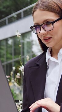 Young businesswoman in formal attire typing on a laptop outdoors in a city park, with a modern glass office building in the background. Concept of business, remote work, productivity, career