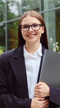 Portrait of a young businesswoman in a professional business suit standing outdoors with a laptop, looking at the camera and smiling against the background of a modern glass office building. Concept