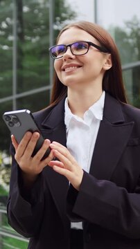 Young woman in formal business attire holding a mobile phone and looking to the side against the background of a modern glass office building. Business communication, career and technology concept