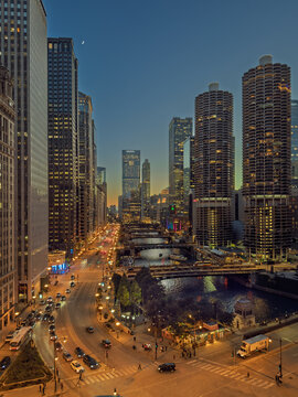 CHICAGO, IL, JUN 18, 2026: Dusk over the Chicago River with Marina City, Salesforce Tower and a crescent moon, glowing bridges, a bus and truck at a busy intersection, and a lit riverside kiosk