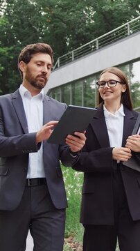Young businesswoman and businessman walking through a city park, discussing work while looking at a tablet screen held by the man. Slow motion