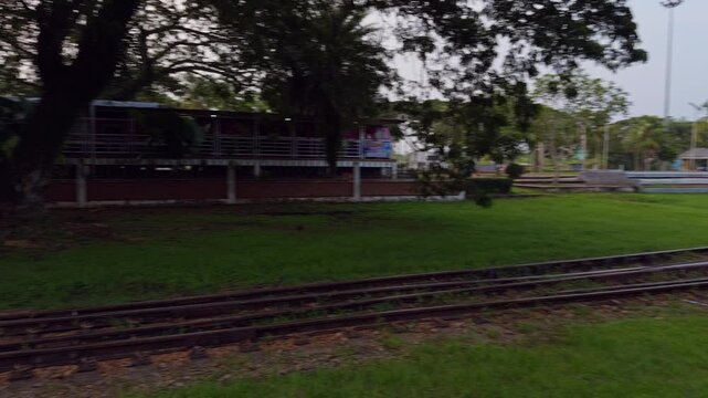 Railway Tracks Extend Beside an Empty Station Platform, with Gravel Beds, Green Grass, Palm Trees and Shaded Buildings, Showing Rail Transport Infrastructure in a Leafy Outdoor Setting