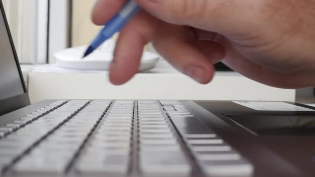 Hand Holding Blue Pen Writing on Laptop Keyboard. Typing on Laptop Keyboard Closeup Detail.