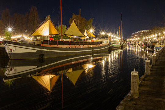 View of a traditional sailing boat moored in a canal at night, with illuminated sails reflecting on the calm water and streetlights lining the quay in Amersfoort, Utrecht, Netherlands.