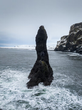 View of Reynisdrangar sea stack rising from the turbulent North Atlantic ocean with snow-covered cliffs in the background, Iceland.