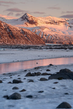 View of snow-covered mountains reflecting in a frozen glacial landscape under the warm light of sunset in Iceland.
