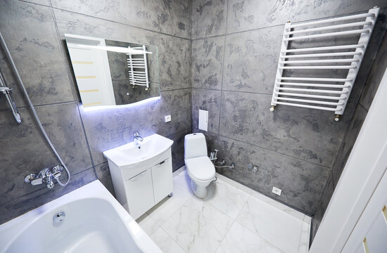 Modern bathroom interior with sink cabinet, toilet and heated towel rail. Stylish washroom with dark textured walls, marble floor tiles, backlit mirror and bathtub edge