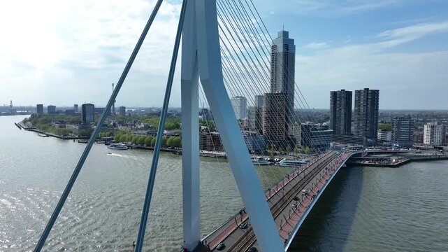 Aerial drone view of the iconic Erasmus Bridge spanning the Nieuwe Maas River in Rotterdam, Netherlands, with modern skyscrapers, boats and urban waterfront scenery.